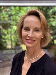 A woman with short blonde hair, wearing a black top, pearl necklace, and earrings, smiles at the camera in an outdoor setting with greenery behind her. This is Jordan Vaughan Smith.
