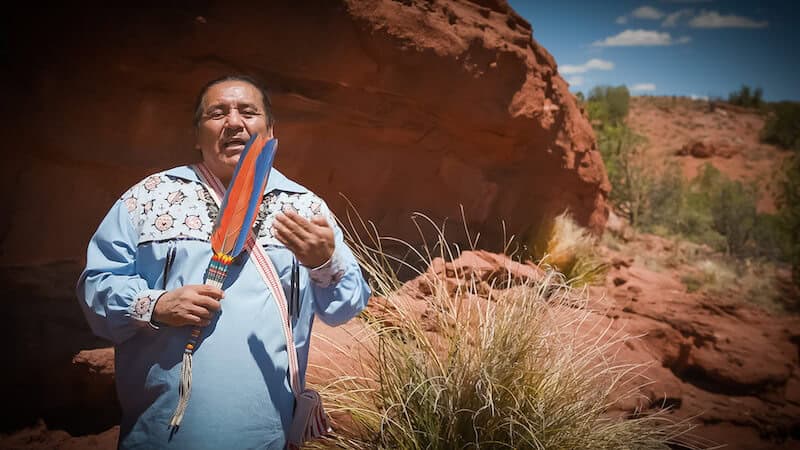 Brophy Toledo stands before red rocks holding an Indigenous piece made of beadwork and feathers