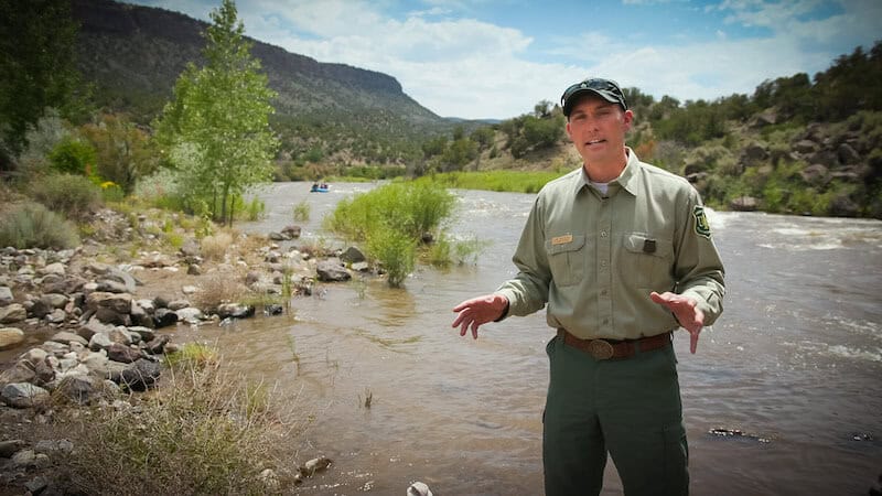 Carl Malcom explains something along the Chama river with a mesa in the background