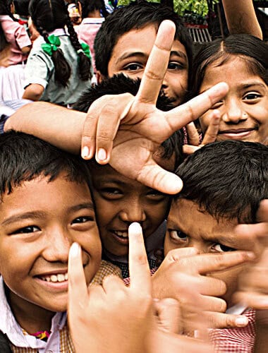 A group of smiling children pose closely together, some making playful hand signs and looking directly at the camera.