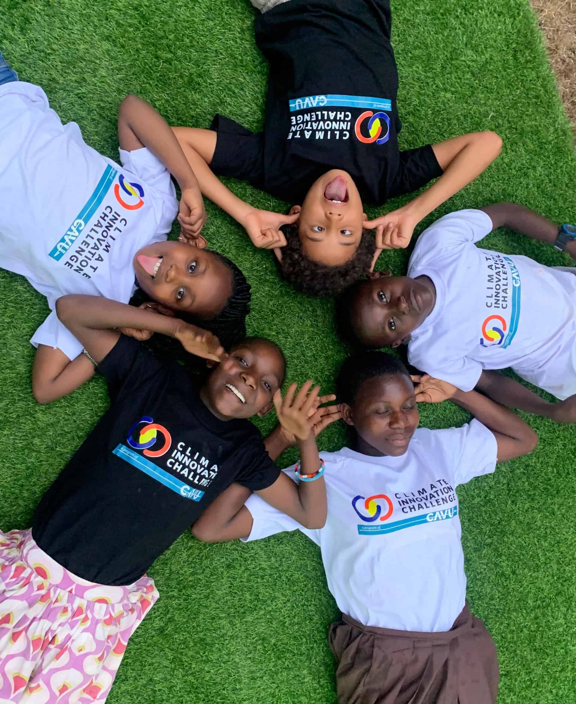 Five children lie on grass in a circle with heads together, smiling and making playful faces. They wear matching “Climate Knowledge Challenge” t-shirts in black and white.
