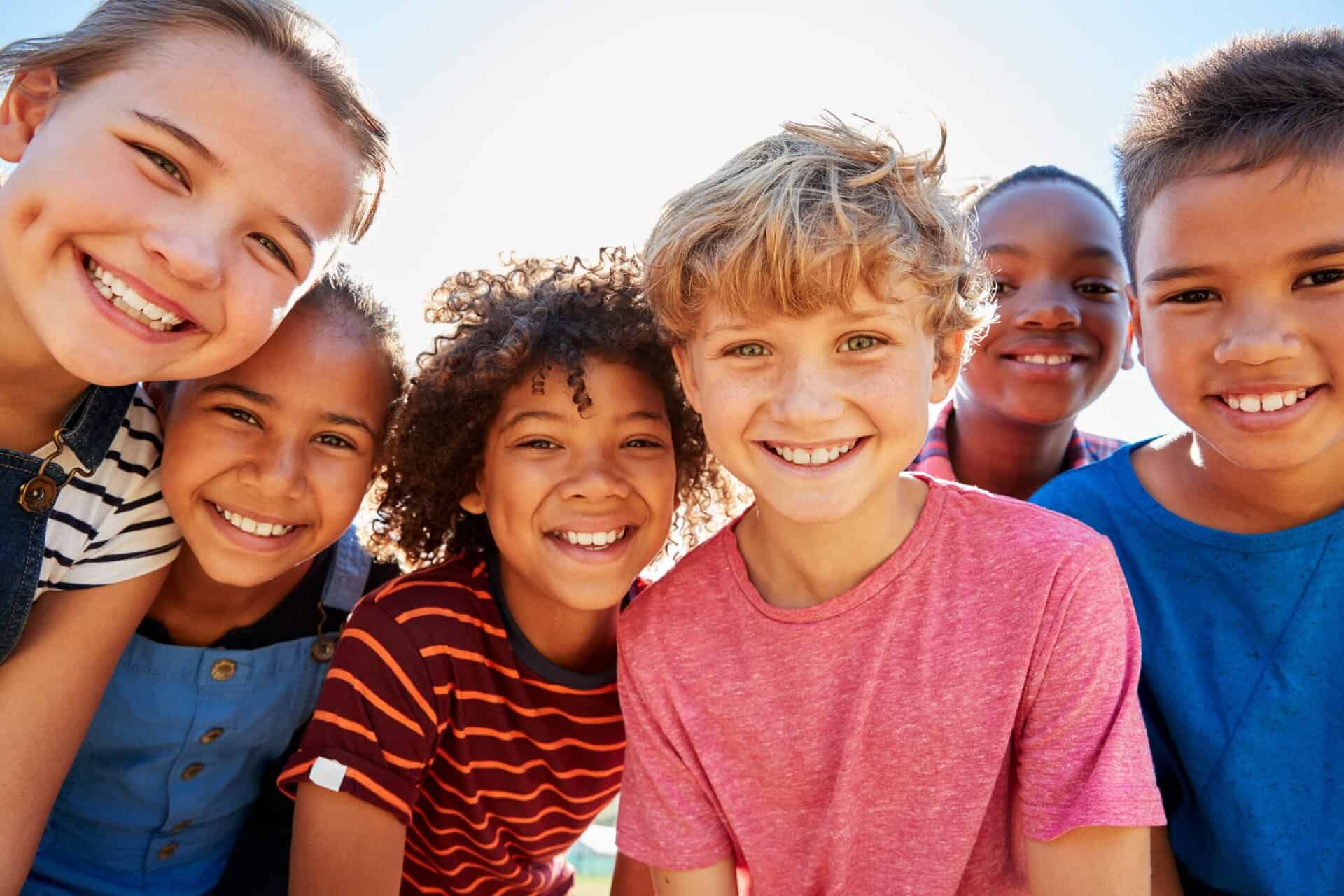 Six smiling children stand close together outdoors, looking at the camera, with sunlight in the background.
