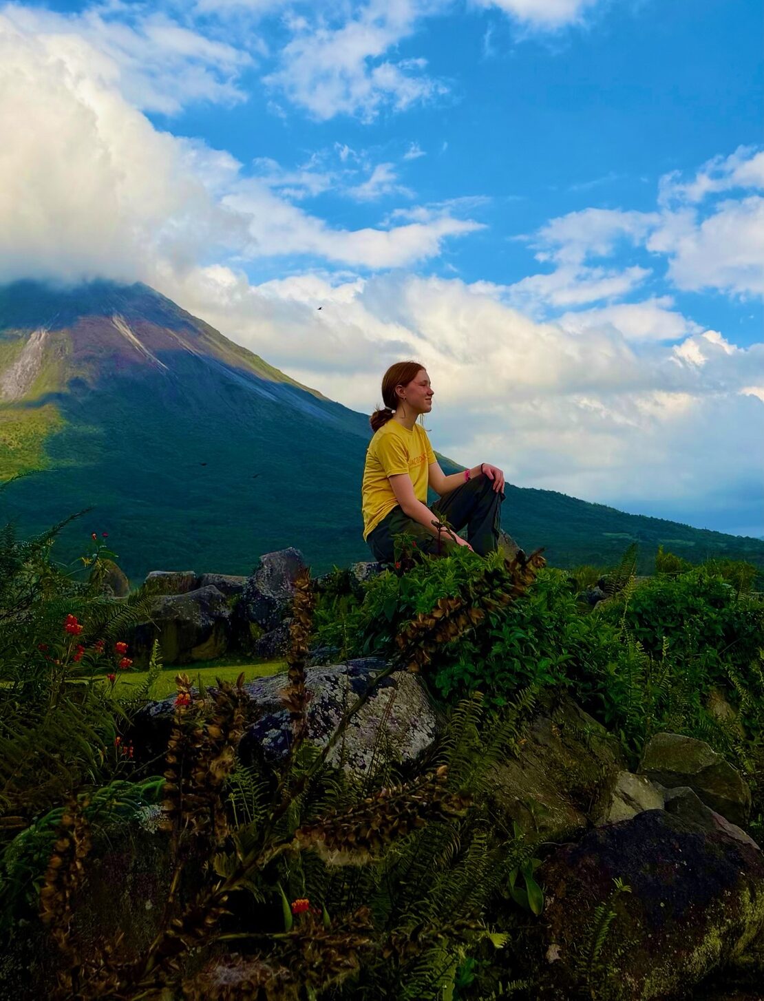 A person in a yellow shirt sits on rocks in front of Bleakley Shettle, a green mountain with a cloud-covered peak under a blue sky with scattered clouds.
