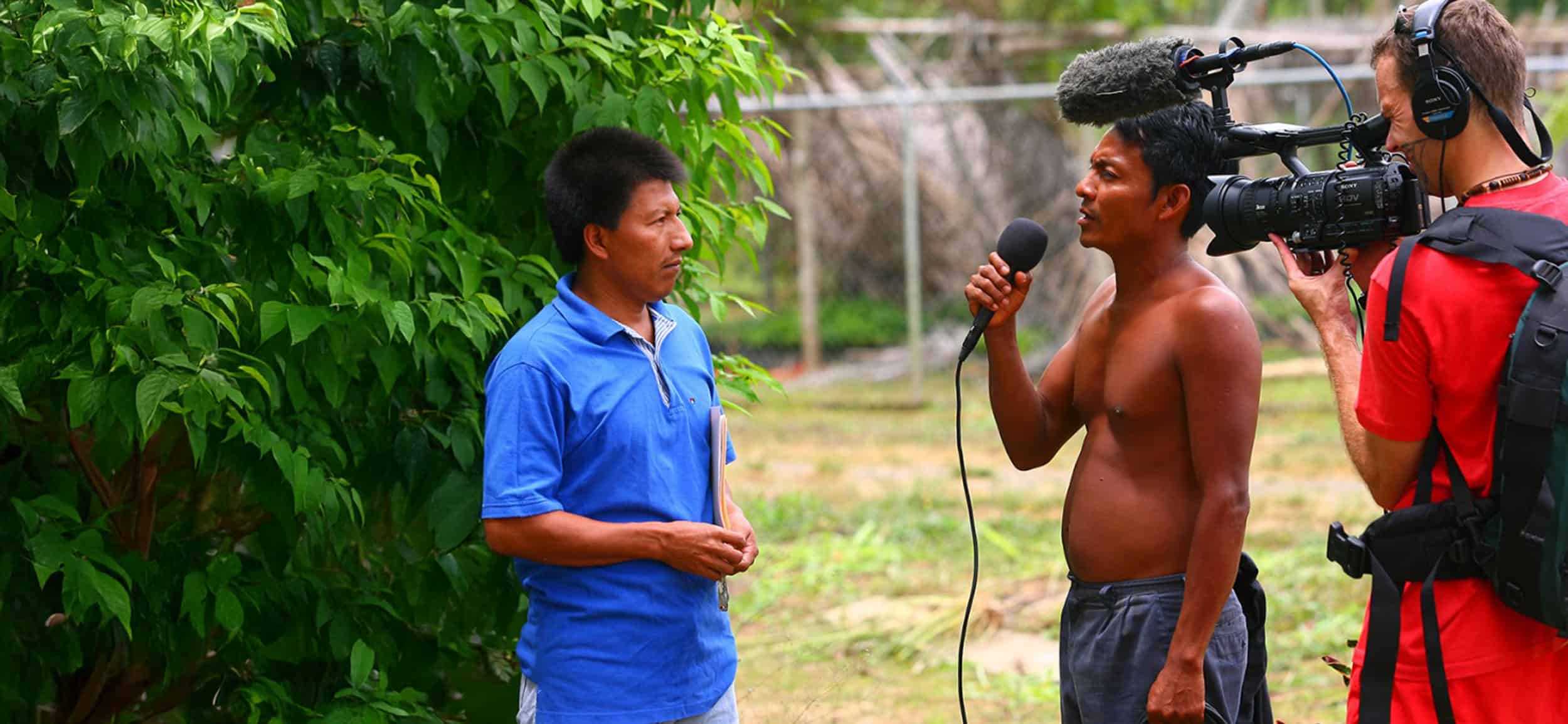 A shirtless man holds a microphone and interviews another man in a blue shirt while a cameraman films them outdoors near green foliage.