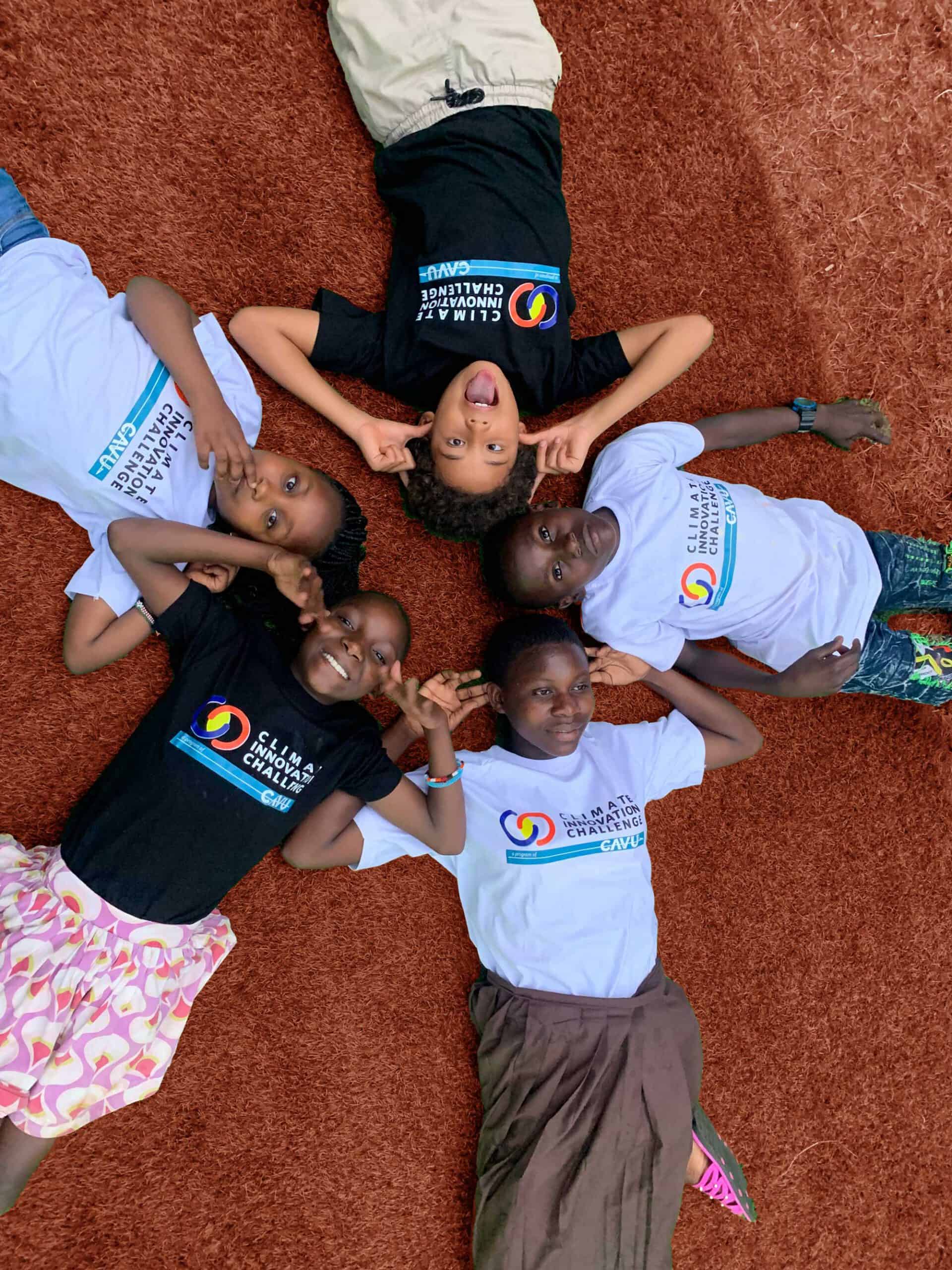 Five children lie on their backs in a circle on reddish-brown ground, touching heads, wearing black or white t-shirts with colorful logos and text.