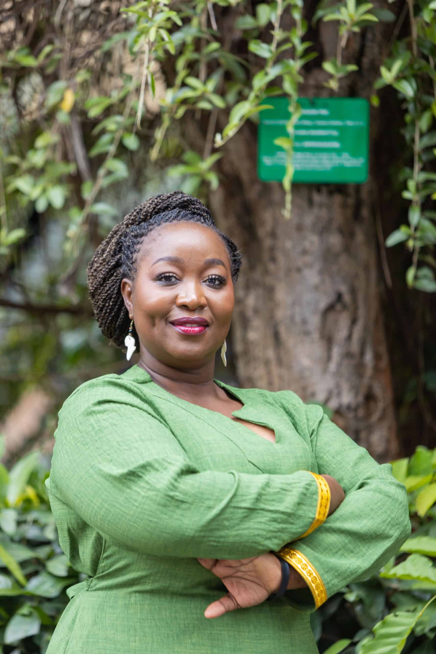 Lorna Wambayi, in a green dress, stands outdoors with arms crossed, smiling warmly amid lush greenery and a tree in the background.