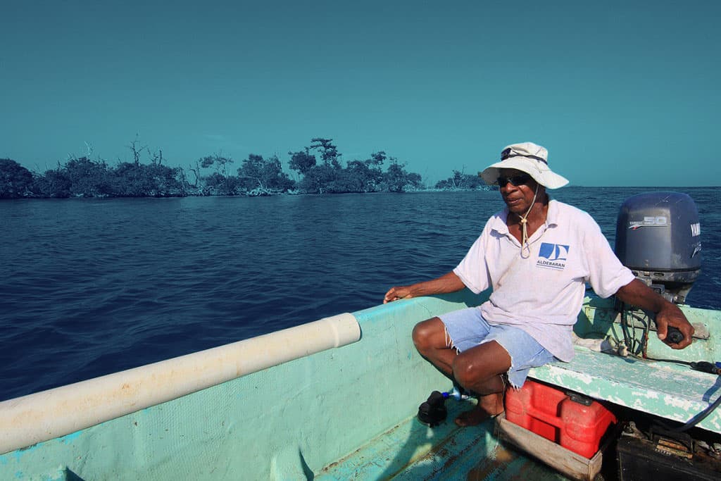 A person wearing a hat and sunglasses sits in a small motorboat on the water, with a distant shoreline and trees in the background.