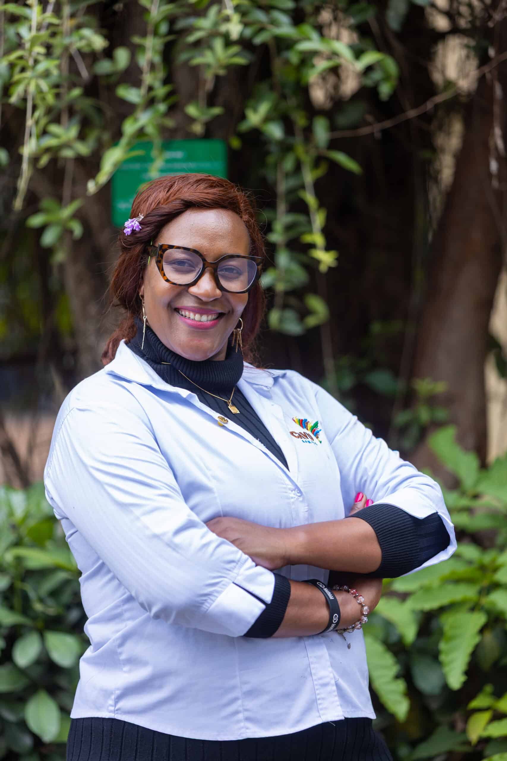 Lorna Wambayi, wearing glasses and a white lab coat, stands outdoors with arms crossed, smiling at the camera. Lush green plants and trees form the backdrop.