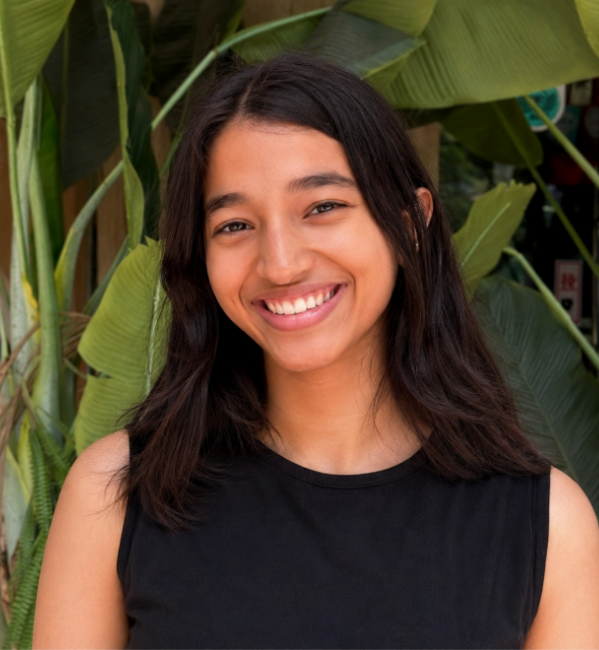 Young woman with long dark hair, wearing a sleeveless black top, smiling in front of large green tropical plants—Shanaaya Chaudhary radiates effortless charm and natural beauty.
