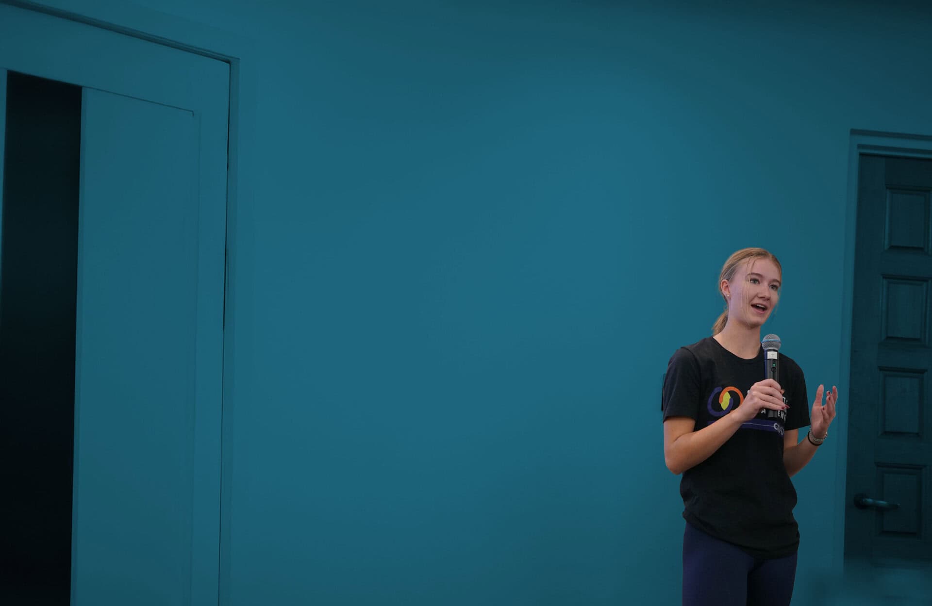 A person holding a microphone speaks while standing near a closed door, against a plain blue wall background.