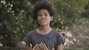 A person with curly hair speaks outdoors, gesturing with their hands, wearing a dark t-shirt and a small round device clipped to their shirt. Green foliage is visible in the background.