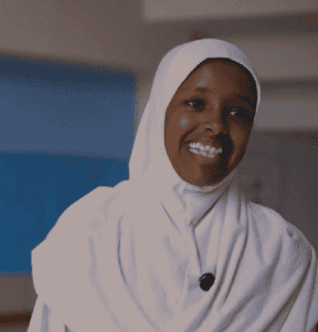 A woman wearing a white hijab smiles while standing indoors in a softly lit room.
