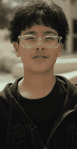 A young person with short, dark hair and glasses is standing outdoors, wearing a black shirt and zip-up jacket, looking toward the camera.