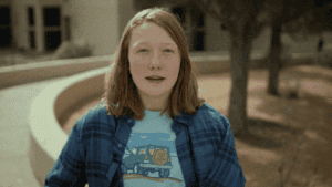 A person with shoulder-length hair stands outdoors, wearing a blue plaid shirt over a graphic tee with a pickup truck illustration. Trees and a building are visible in the background.