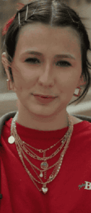 A person with hair clips, multiple chain necklaces, square earrings, and a red shirt looks at the camera.