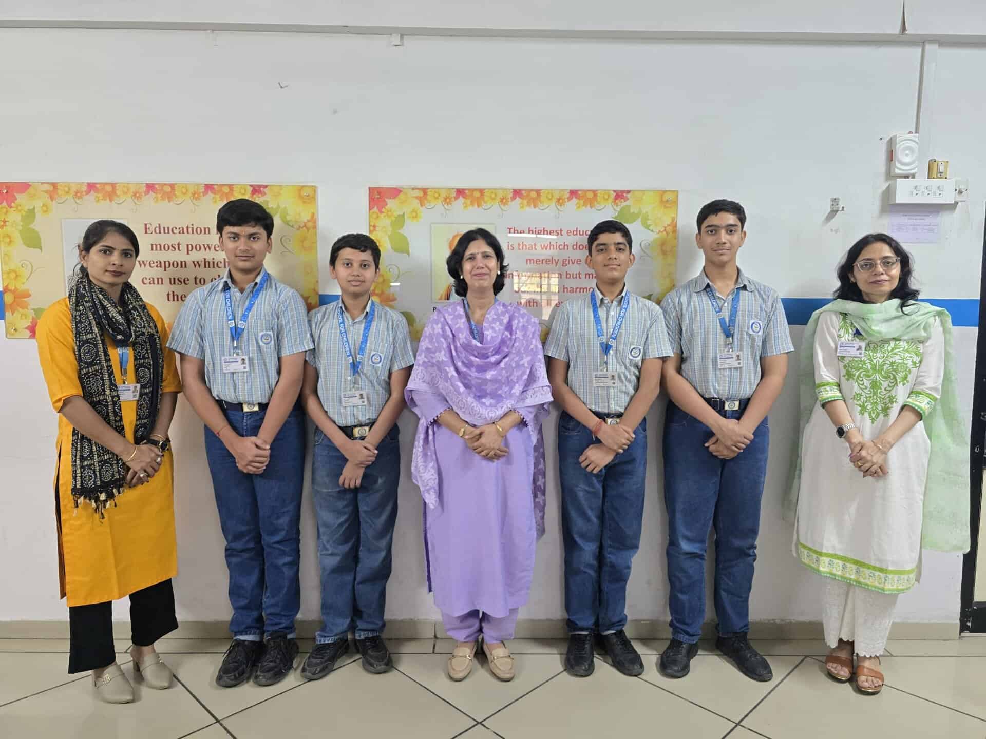 A group of four students in blue uniforms stand between two women in traditional attire at Zebar School, all facing the camera inside a school hallway, reflecting the spirit of innovation and community at CIC.