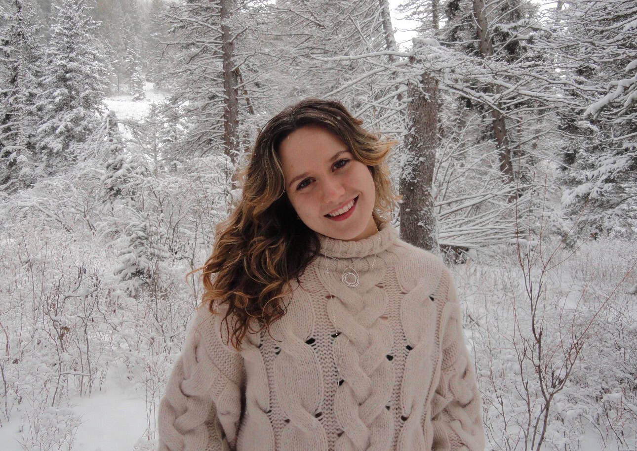 Lucy Sullivan, dressed in a beige cable-knit sweater, stands outdoors in a snowy forest, smiling at the camera. Snow blankets the trees and ground behind her.