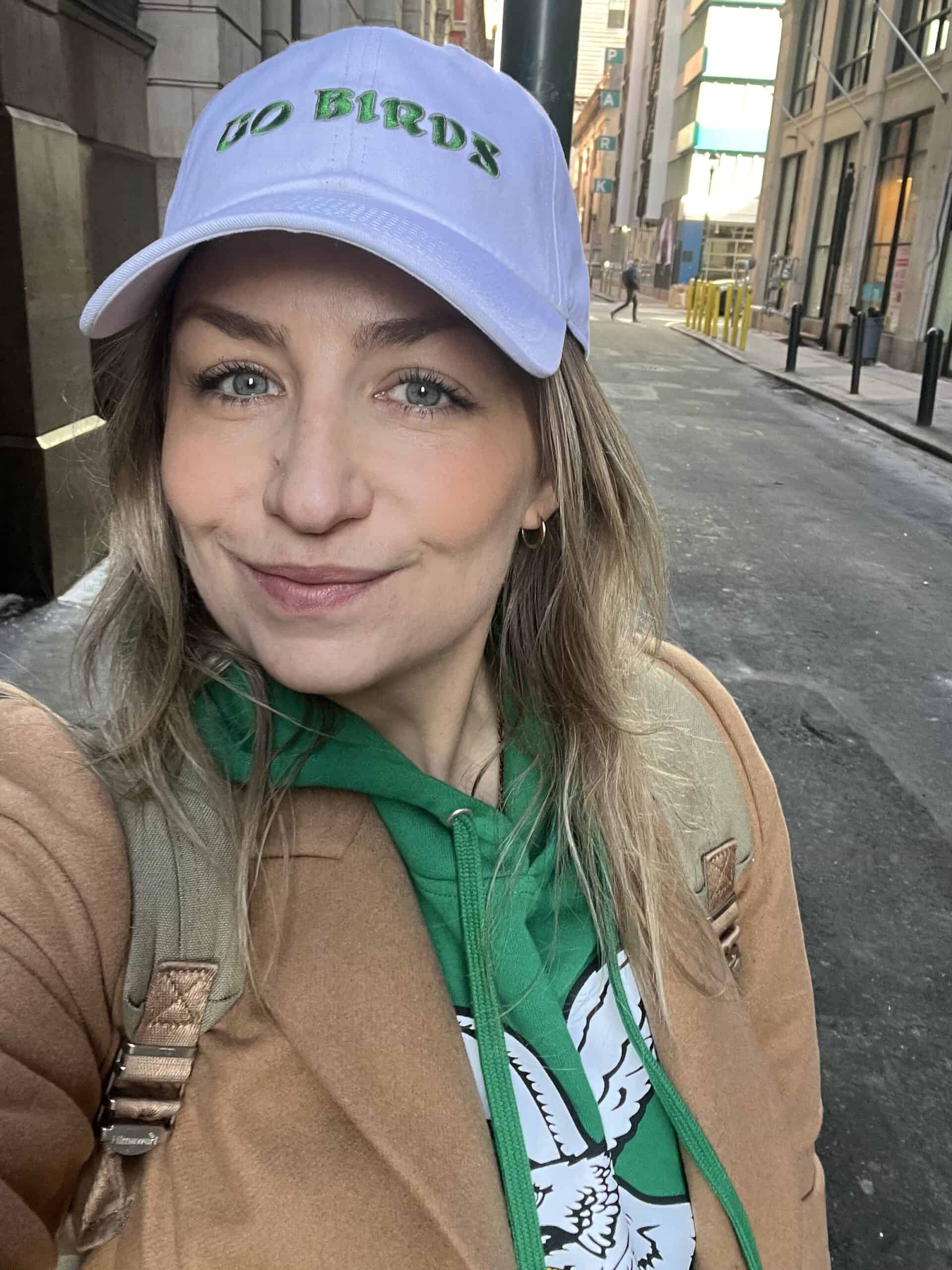 Kimberly Baker, wearing a white Go Birds cap, green hoodie with a bird graphic, and tan coat, stands on a city street taking a selfie.