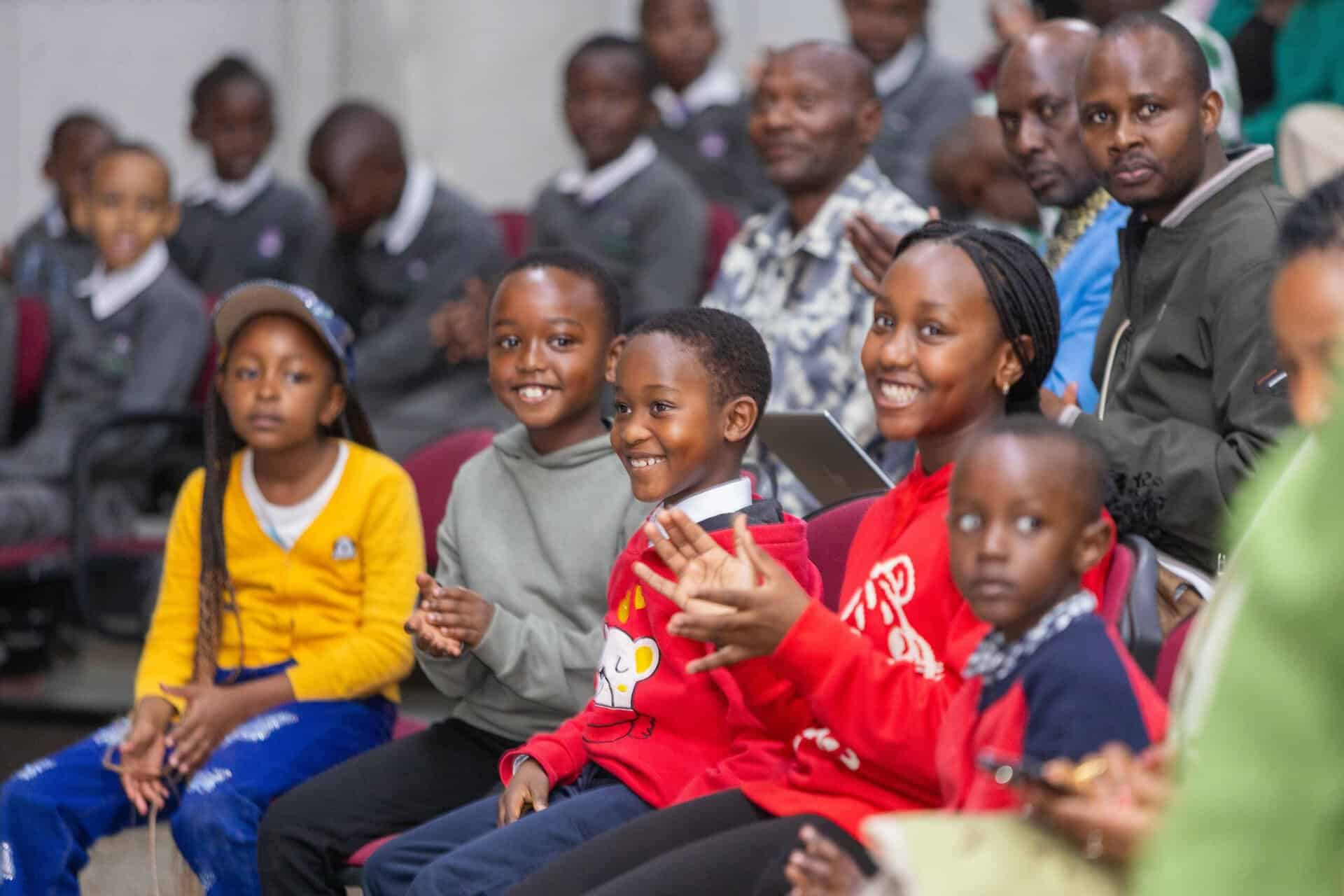 A group of children and adults sit together indoors, smiling and clapping while facing forward, with some wearing colorful sweaters—a joyful moment featured in the July 2026 edition of the Hope in Action Newsletter.
