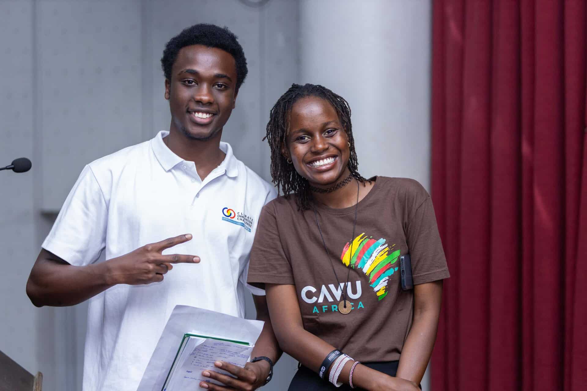 Two young adults smiling at the camera; one holds papers and flashes a peace sign, the other wears a CAVU Africa t-shirt—captured indoors with a red curtain, featured in our July 2026 Hope in Action Newsletter.