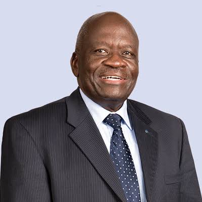 A man wearing a dark pinstripe suit, white shirt, and navy polka dot tie—Francis Okomo-Okello—smiles at the camera against a plain light background.