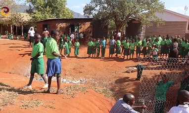 Children in green uniforms gather outside school buildings on a dirt yard, with some adults present and a wire fence in the foreground.