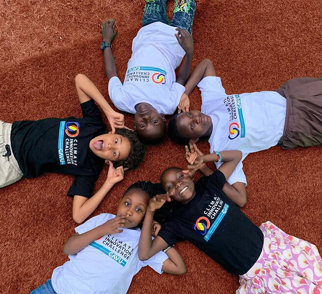 Five children lie on the ground in a circle, heads together, wearing Climate Change Challenge t-shirts and smiling at the camera.