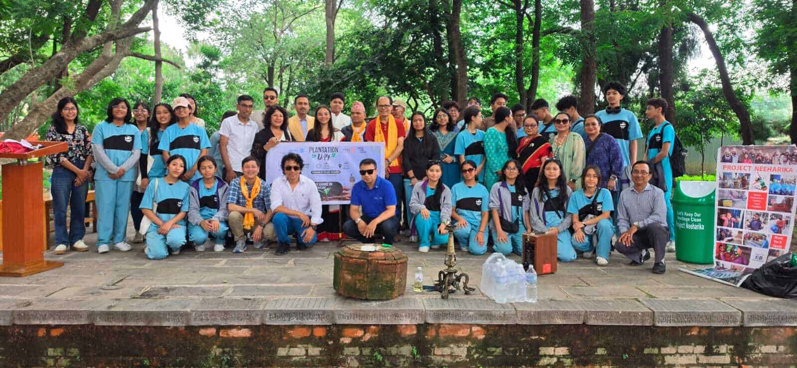 A group of people pose outdoors in front of a display board and banner, surrounded by trees. Most wear matching blue uniforms; some wear casual clothing. United as students build hope, they gather with the tools to build hope featured in the Hope in Action Newsletter.