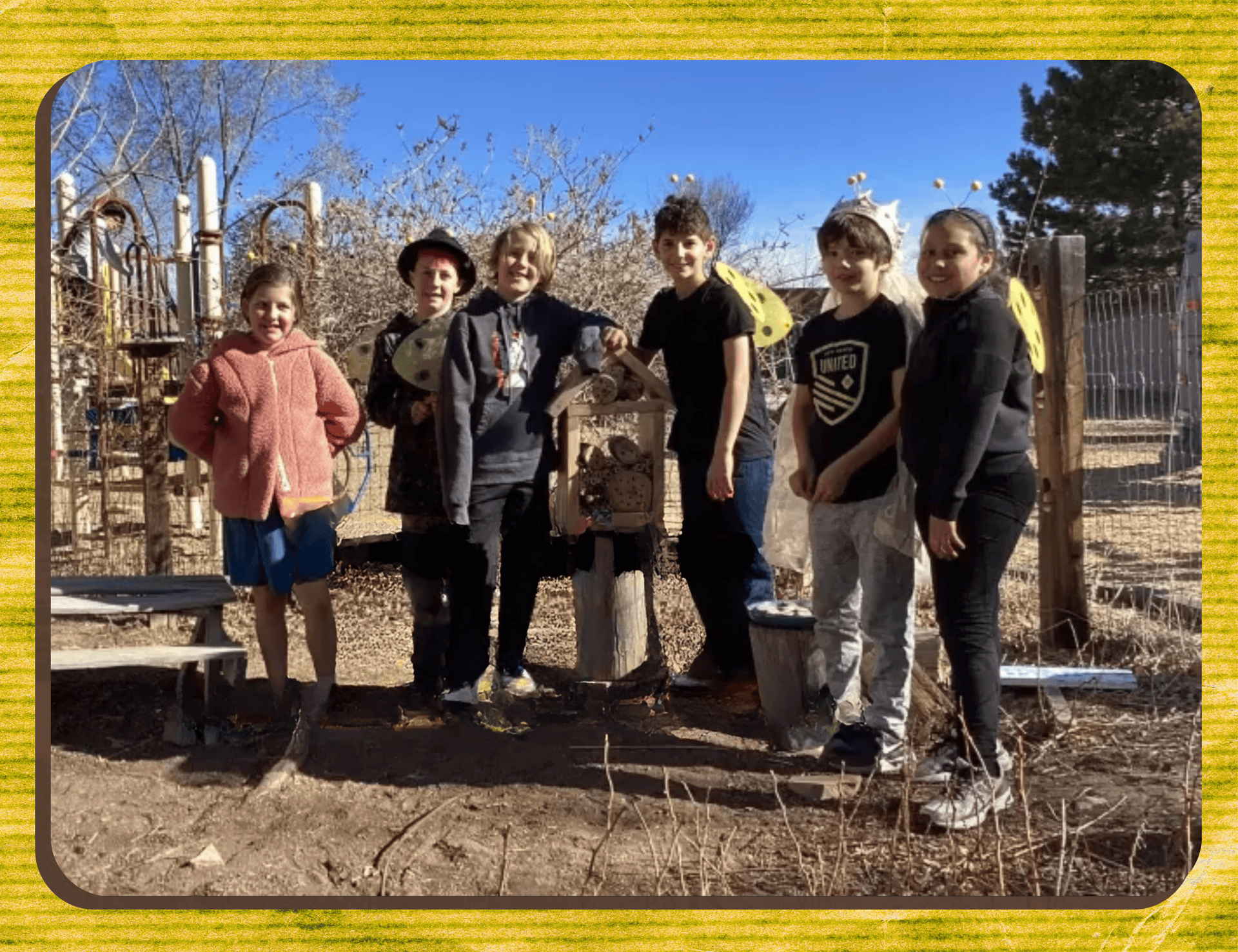 Six children stand outdoors near a bench and playground equipment, some wearing yellow wings and headbands, on a sunny day with bare trees in the background—demonstrating how students build hope through playful connection and creativity.