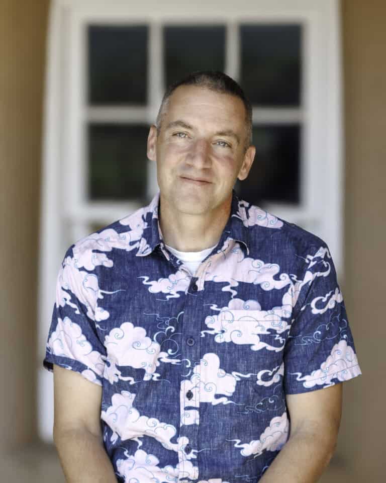 A man with short hair wearing a blue shirt with pink cloud patterns sits in front of a blurred doorway, looking at the camera with a slight smile—embodying the spirit of the Hope in Action Newsletter, where students build hope through everyday moments.