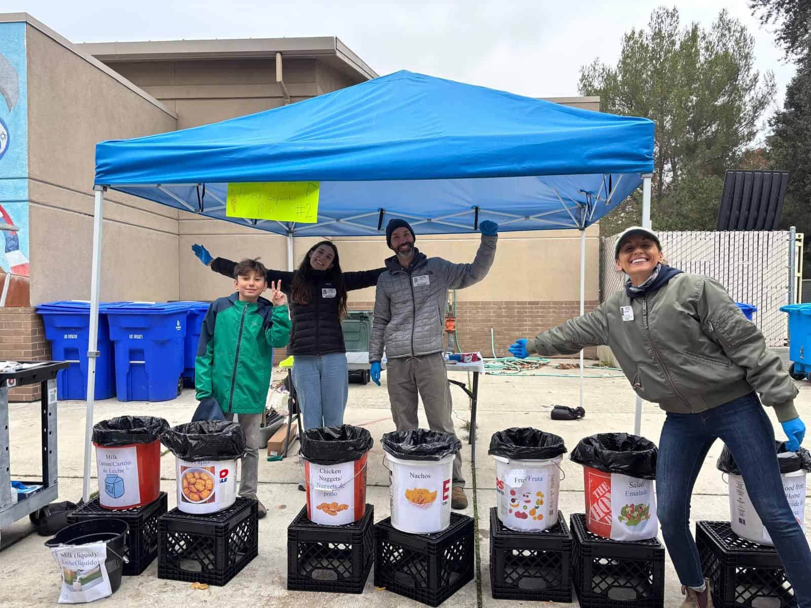 Four people stand under a blue canopy next to bins labeled for different types of food waste, with recycling bins and buildings in the background—an uplifting scene of climate inspiration in action.