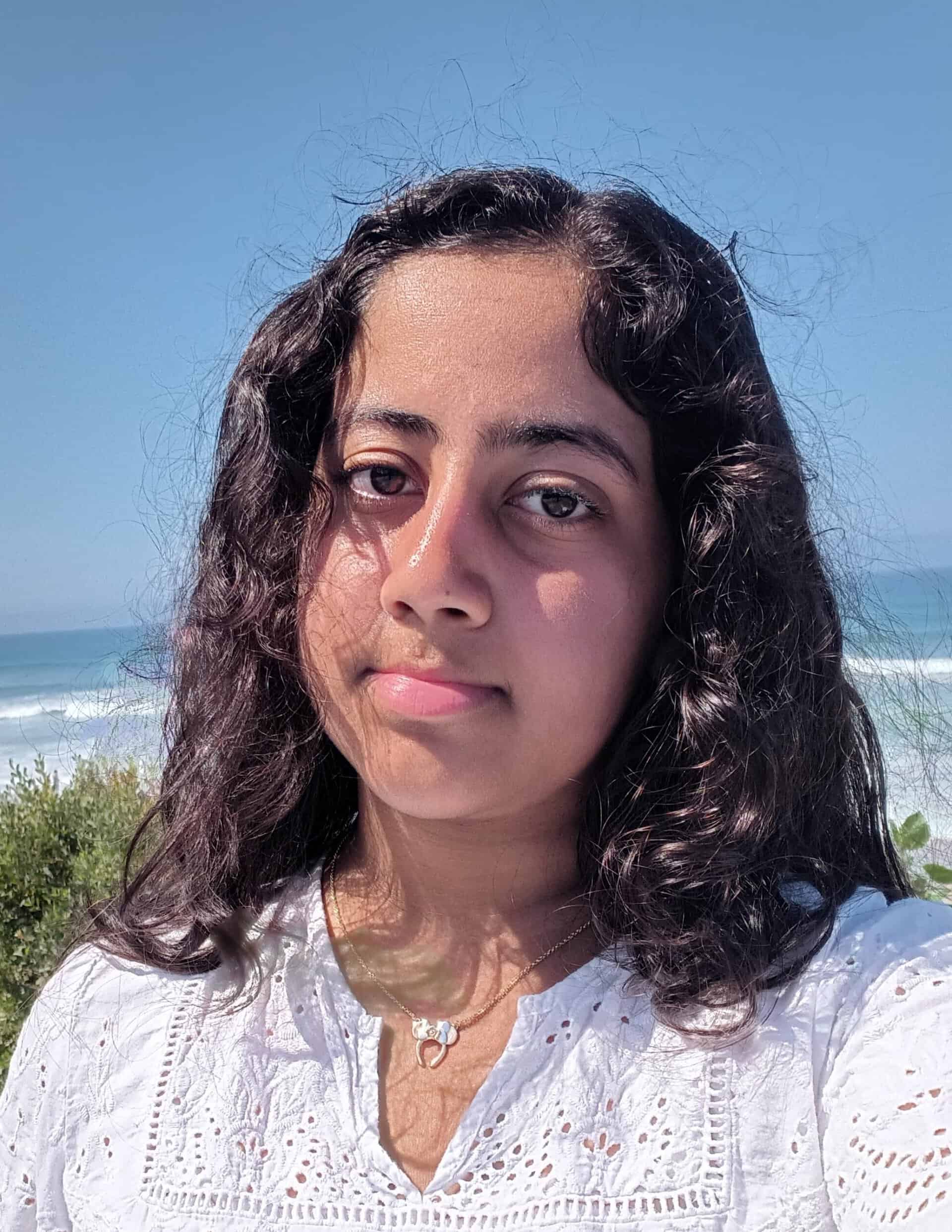 A person with curly hair and a white top stands outdoors near the ocean on a sunny day, with blue sky and waves in the background.
