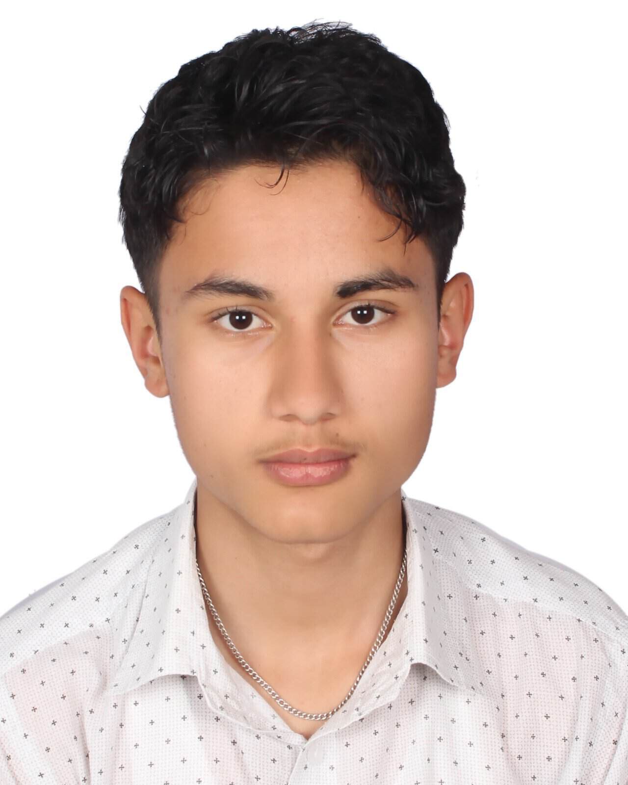 A young man with short, dark hair wearing a white, dotted shirt and a silver chain necklace poses against a plain white background.