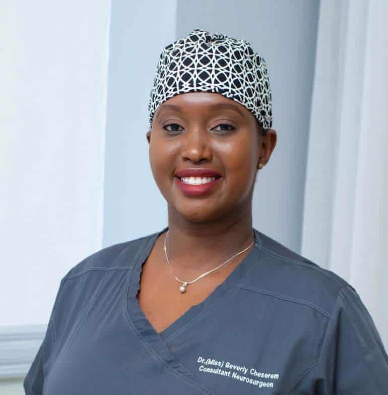 A woman in gray medical scrubs and a patterned surgical cap smiles at the camera. Her scrubs have text that reads Consultant Neurosurgeon, and she is identified as Dr. Beverely Cheserem, FRCS.
