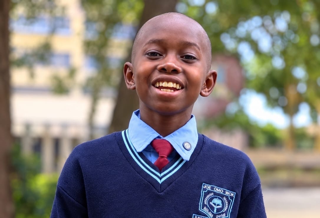 Ian Nasio Wycliffe, a young student, smiles while wearing a school uniform consisting of a light blue collared shirt, a dark maroon tie, and a dark blue V-neck sweater with a school crest.
