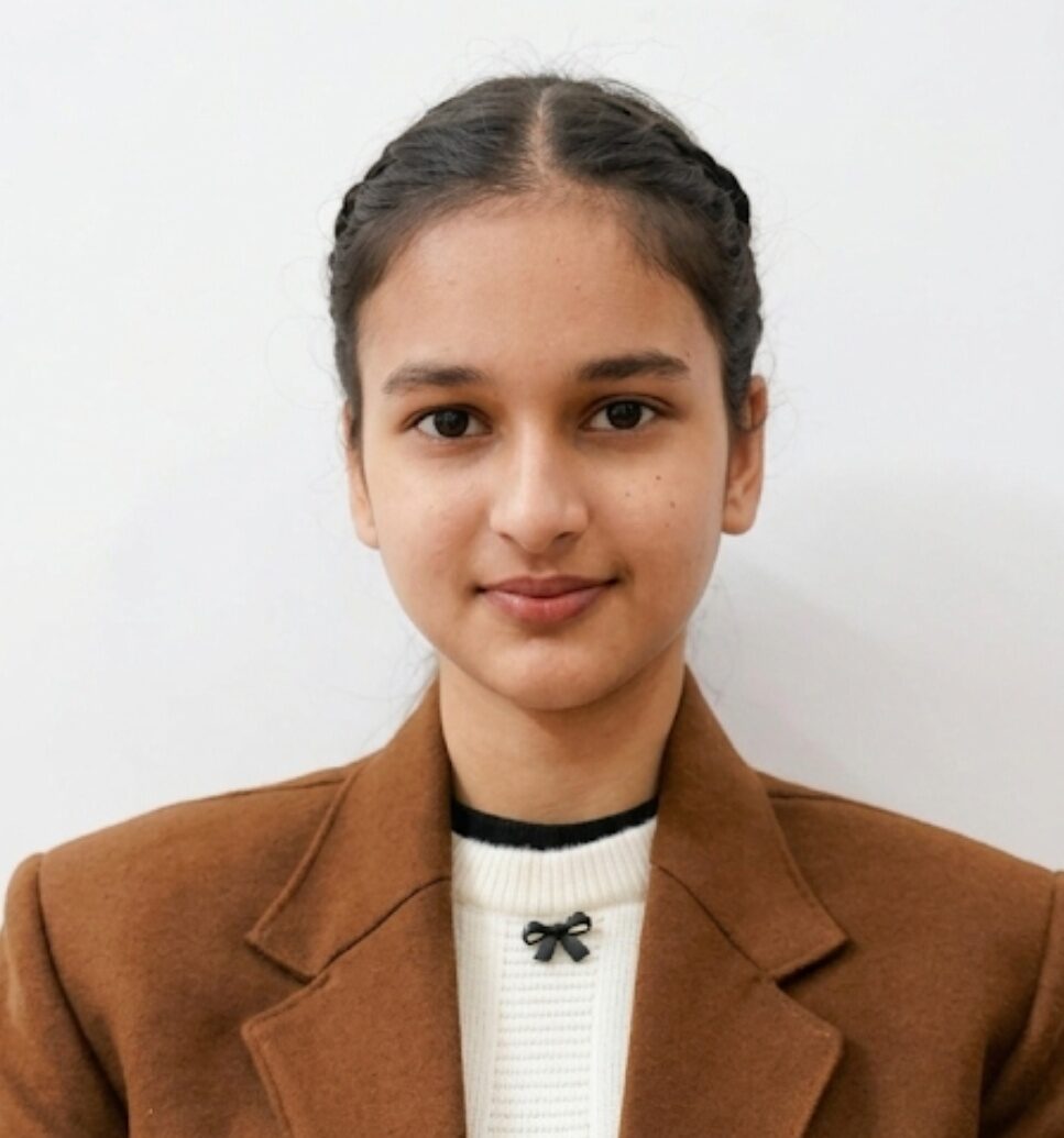 Tanishreet Kaur, a young person with braided hair, is wearing a brown blazer over a white sweater with black bows, standing against a plain white background.