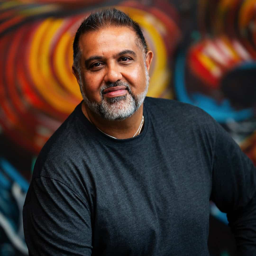 Middle-aged man with a graying beard and short hair, wearing a long-sleeved dark gray shirt against a colorful, abstract background.
